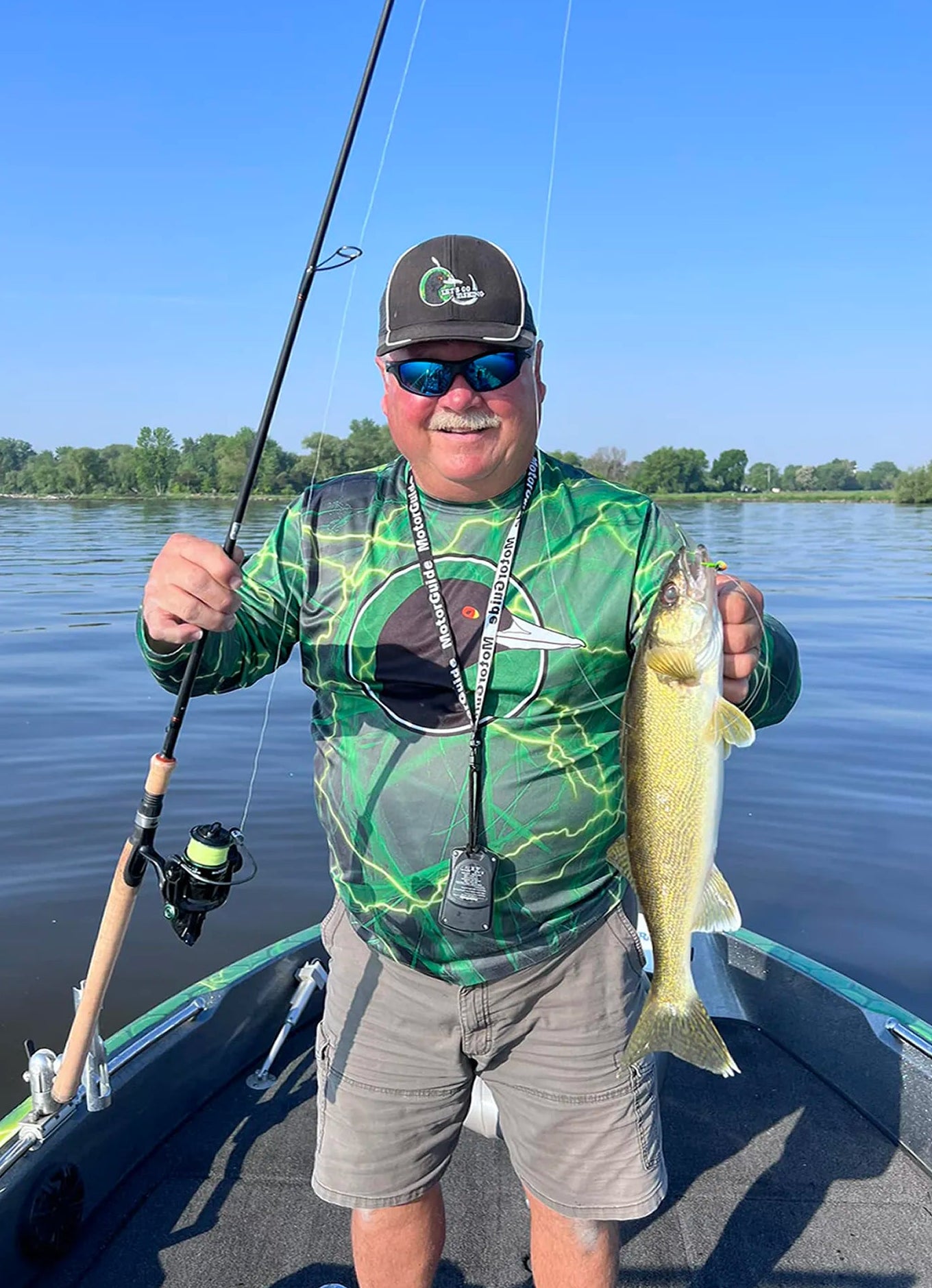 Man holding a fish on a boat with a fishing rod, surrounded by water and trees.