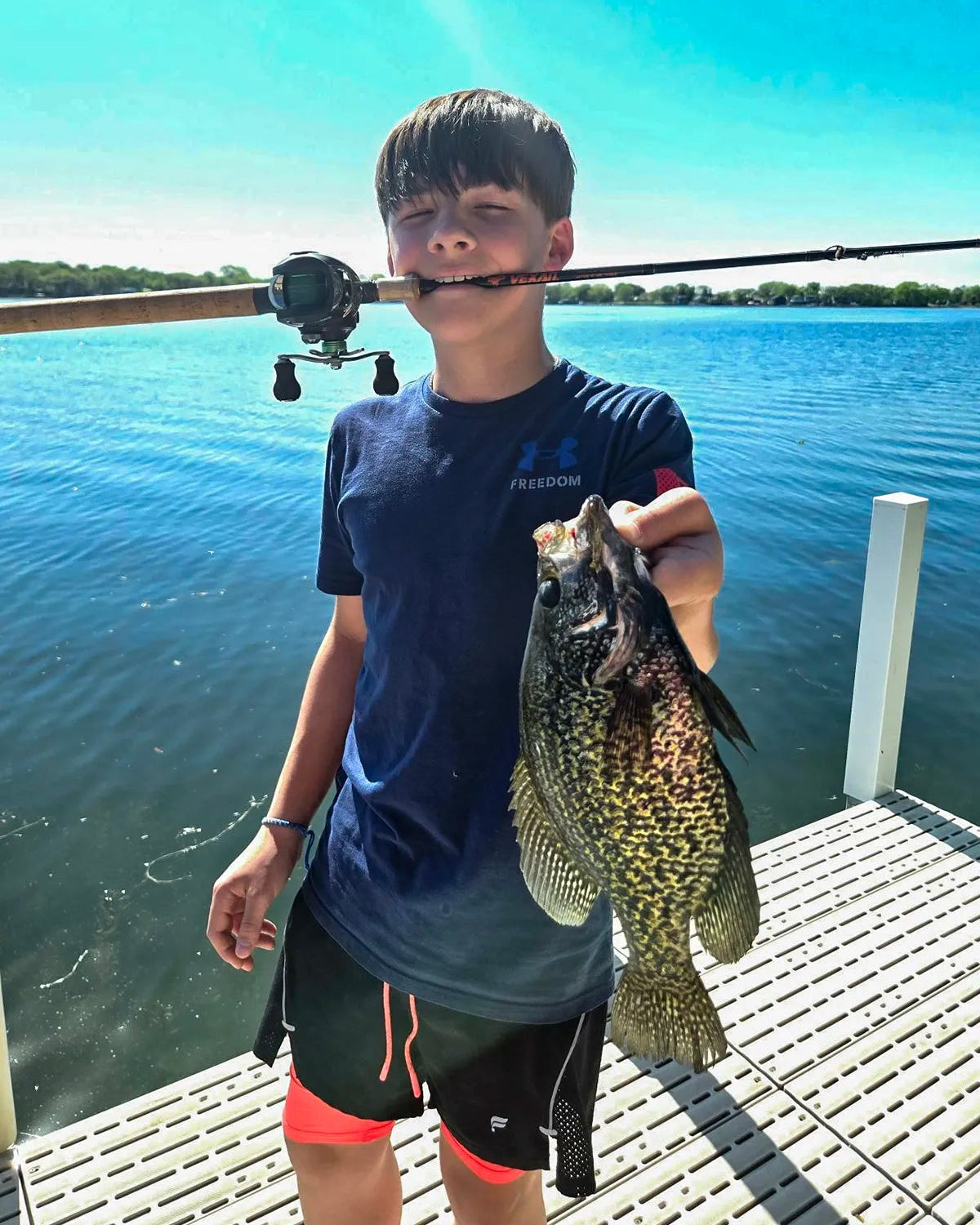 Boy holding a fish on a dock with a clear blue sky and water background